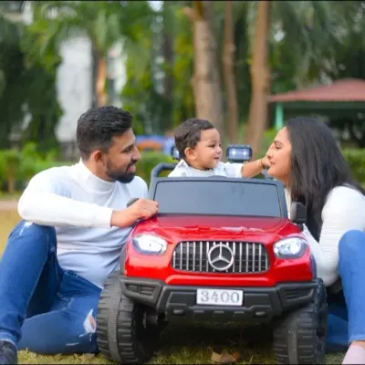 A candid moment from a family photoshoot in the park. The parents share a loving moment with their child, who is having a blast in his toy car.