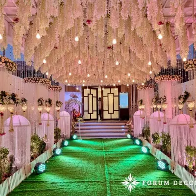 This dreamy pastel walkway creates a magical path for guests. The ceiling is a dense canopy of hanging white wisteria and light bulbs, creating a starry night effect. The green turf path is lined with draped pillars and traditional hangings, making the walk to the main event an experience in itself.