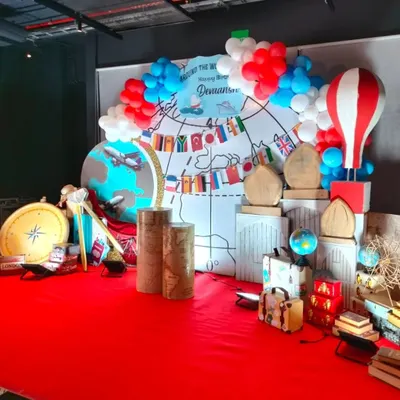 A full stage setup for an "Around the World" birthday theme, featuring a world map backdrop, globes, suitcases, and a hot air balloon prop.