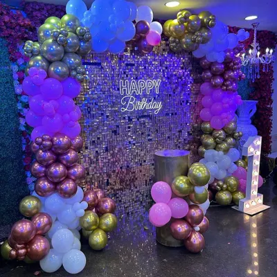A glamorous birthday setup with a gold shimmer wall as the centerpiece. The decor is accented with purple, pink, and gold balloons, a 'Happy Birthday' neon sign, and a light-up number '1'.