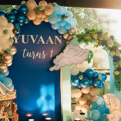 A beautiful royal blue themed first birthday for "Yuvaan," featuring a large blue backdrop personalized with his name. The decor includes a charming carousel prop and an elegant balloon arch in shades of blue, white, and silver, creating a sophisticated and celebratory atmosphere.