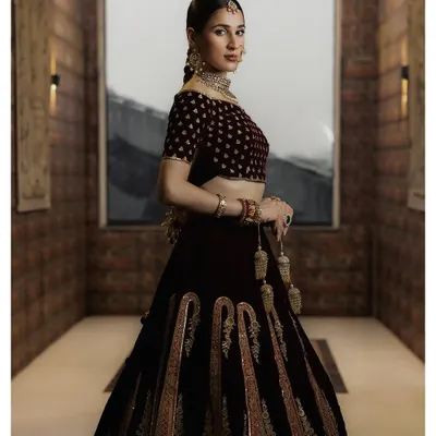 A full-length portrait showcasing a magnificent velvet lehenga with detailed embroidery. This shot captures the entire silhouette and the grandeur of the bridal outfit within a well-composed architectural setting.