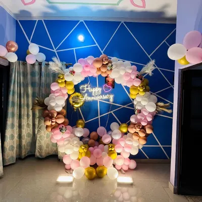 This indoor anniversary decoration features a full circular balloon arch in pastel pink, white, and gold tones. A 'Happy Anniversary' neon sign hangs in the center, creating a beautiful focal point in the room.