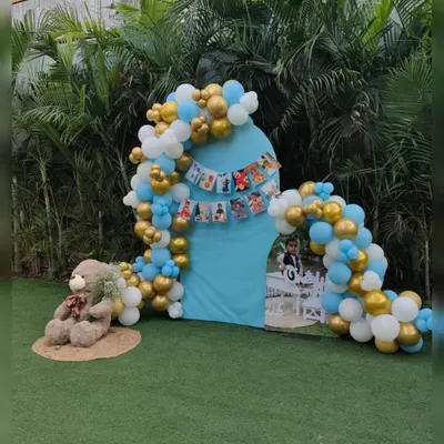 A beautiful outdoor setup for a first birthday celebration, featuring a teddy bear theme with a blue, white, and gold balloon arch.