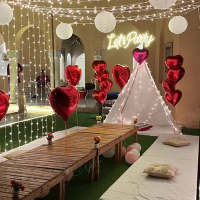 A beautiful cabana tent setup for an outdoor party. This arrangement features low seating with cushions, a 'Let's Party' neon sign, heart-shaped balloons, and enchanting fairy lights, creating a cozy and celebratory vibe.