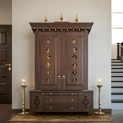 This freestanding pooja cabinet is a masterpiece of traditional craftsmanship. Made from dark wood, it features doors with small, bell-shaped cutouts, a decorative top cornice, and ample drawer storage at the base. It stands as a beautiful, self-contained temple within your home.
