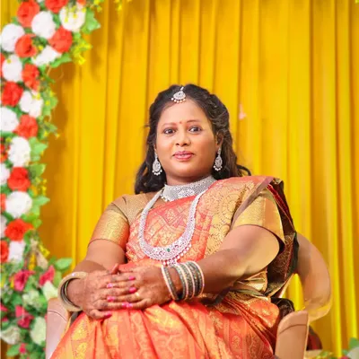 A classic portrait of the bride at her event. Seated against a vibrant yellow and floral backdrop, her elegant saree and jewelry are beautifully highlighted.