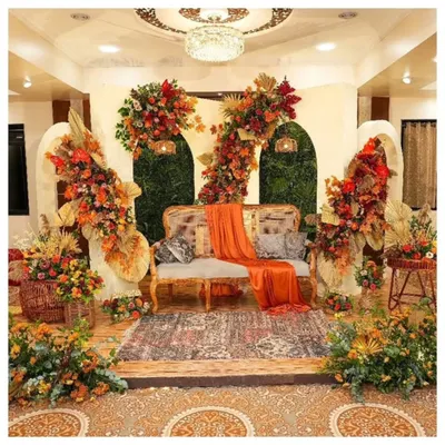 A rich, rustic-themed stage setup perfect for a Haldi or Mehendi ceremony. I used a warm, autumnal color palette with orange and red flowers, dried palm leaves, and pampas grass, all centered around a comfortable cane sofa to create a cozy and photogenic space.