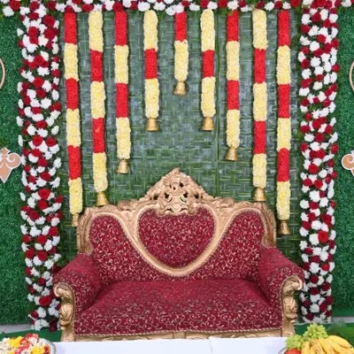 A traditional Seemantha backdrop made with a woven coconut leaf mat. The stage is decorated with red and white floral borders and hanging garlands of yellow chrysanthemums and bells.