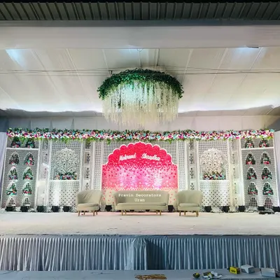 This indoor wedding stage combines royal and floral elements. We used white jali panels, a custom pink backdrop with the couple's names, and a stunning floral chandelier with hanging wisteria to create a rich, layered look.
