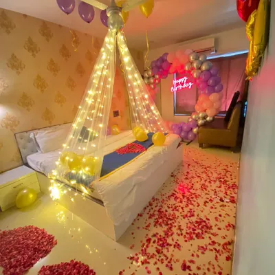 A complete romantic room decoration for a birthday, featuring a fairy light canopy over the bed, a balloon arch, and a floor scattered with rose petals.
