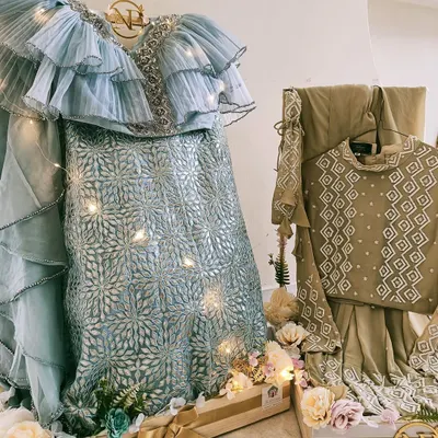 A breathtaking display for a modern bride. This pastel blue ruffled lehenga is presented on a custom wooden stand, adorned with fairy lights and a personalized monogram, creating a stunning focal point for the trousseau.