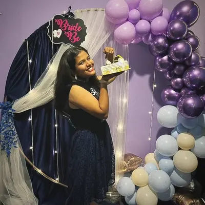 The bride-to-be holding her cake in front of the custom decoration. The setup includes a circular arch with drapes, fairy lights, and a balloon garland in her chosen colors.