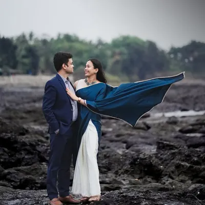 A dramatic shot of a couple on a rocky shore, with the bride's blue saree flowing in the wind. This creates a cinematic and powerful pre-wedding image.