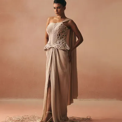 This one-shoulder gown is anchored by a corset featuring intricate cording in a peplum style. It is a piece that balances a strong, sculpted core with the softness of a flowing cape and draped skirt.