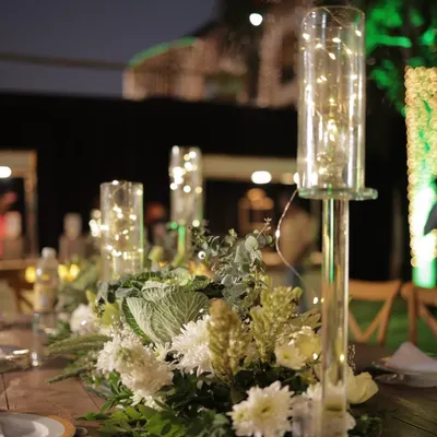 A close-up of a centerpiece featuring fresh white flowers and eucalyptus, with tall glass hurricanes containing fairy lights. This design adds a touch of modern romance and gentle light to the table.