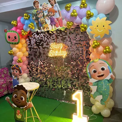 A vibrant Cocomelon theme first birthday party. This setup features a shimmering sequin backdrop, a 'Happy Birthday' neon sign, a colorful balloon arch, and multiple character cutouts from the popular show.