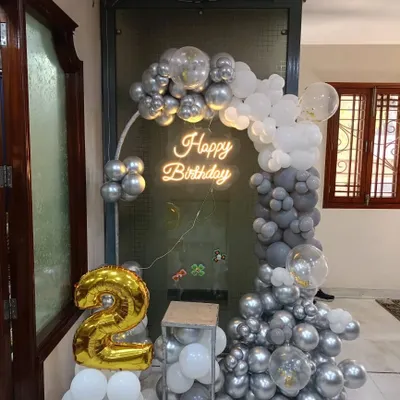 A chic and modern birthday setup for a home. The half-arch of grey and white balloons is accented by a "Happy Birthday" neon sign and a large gold foil number '2'.