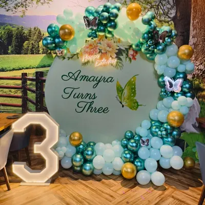 A beautiful butterfly theme for a third birthday. This setup features a custom backdrop, a teal and white organic balloon arch with gold accents, and a large light-up number '3'.