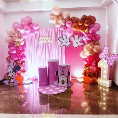 Here is a full view of a Minnie Mouse themed first birthday party. The setup includes a complete balloon arch in shades of pink, a matching pink curtain backdrop, a light-up number '1', and adorable cutouts of Minnie to create a picture-perfect setting.