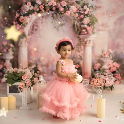 A princess party for a first birthday. This little girl, dressed in a beautiful pink gown, stands in a fairytale setting complete with a floral arch, candles, and a tiny crown.