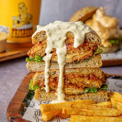 A towering fried chicken sandwich shot for Frisbees Burger Peddler. This image is styled to look indulgent and satisfying, perfect for a new menu launch announcement on social media.