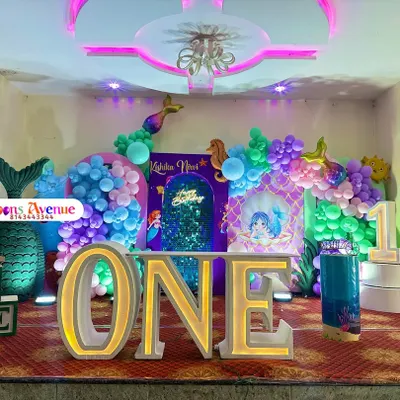An enchanting mermaid-themed first birthday party. This elaborate setup includes a large "ONE" marquee table, a shimmering sequin backdrop, an under-the-sea balloon arch, and numerous mermaid-themed props and cutouts.