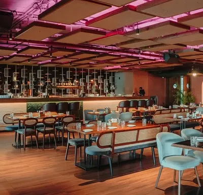 This wide shot captures the dynamic atmosphere of the main dining area and bar. The design uses a mix of seating types, from intimate tables to long benches, all under a ceiling with acoustic panels and dramatic pink lighting.