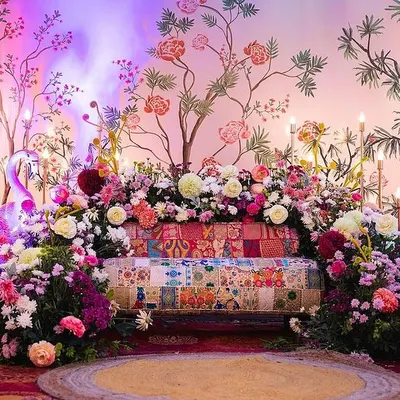 A close-up of a beautiful Mehendi stage with a hand-painted floral backdrop. The decor is enhanced with colorful lighting, a patchwork sofa, and abundant fresh flowers.