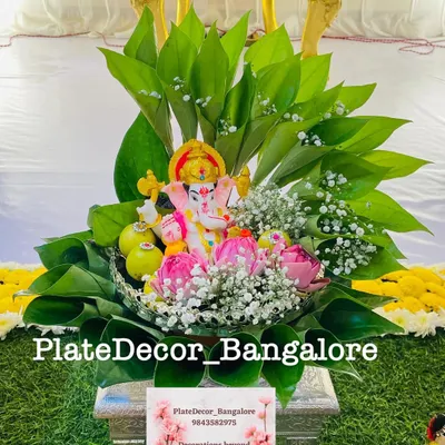 A beautiful pooja platter with a Ganesha idol placed on a bed of betel leaves, surrounded by lotus buds and baby's breath flowers.