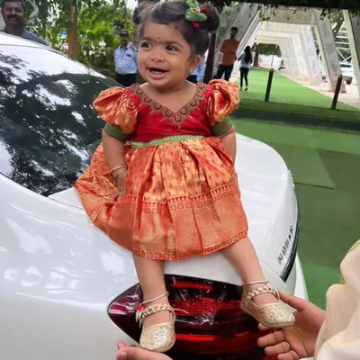 This little princess is all smiles in her custom-designed silk frock. The dress is in a traditional Pattu Pavadai style with a bright red and orange combination and cute puff sleeves.