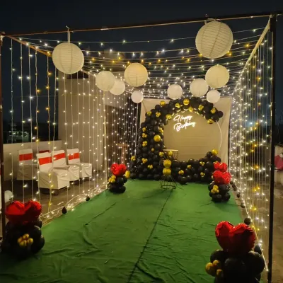 This is a complete birthday party setup I did on a terrace in Greater Noida. I created a magical walkway using curtains of fairy lights and hanging paper lanterns, leading to a stylish black and gold balloon arch.