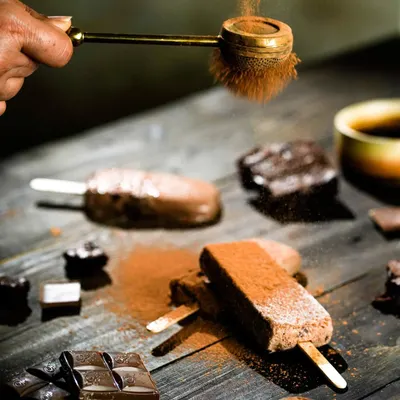 An action shot capturing cocoa powder being dusted over rich, chocolate-flavored popsicles. The dark, moody styling and dynamic motion make this dessert look incredibly indulgent.