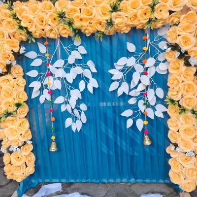 A modern and vibrant pooja backdrop featuring a frame of yellow artificial roses against a blue curtain. The hanging white leaf elements and small bells add a creative touch to the festive decoration.
