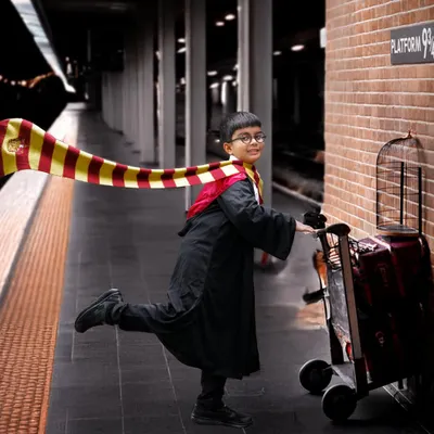 A magical Harry Potter themed photoshoot featuring a young boy in a Gryffindor robe pushing a luggage trolley towards a Platform 9¾ sign on a brick wall.