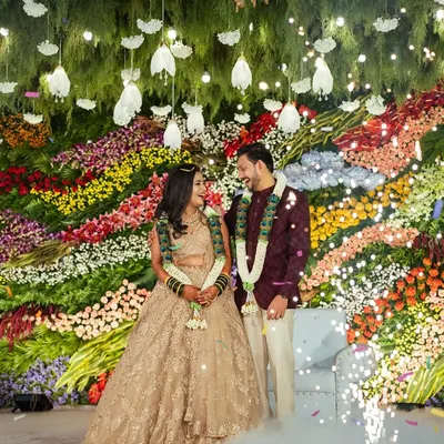 A stunning shot of a couple on their reception stage, set against a massive, vibrant wall of fresh, multi-colored flowers. The falling confetti adds a touch of magic to the moment.