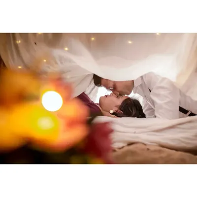 A dreamy and intimate pre-wedding photo, with the couple under a canopy of sheer fabric and fairy lights, creating a magical atmosphere.