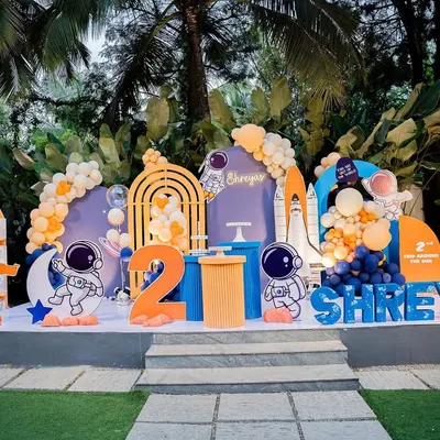 A complete outdoor stage setup for a space-themed second birthday. It features astronaut cutouts, a rocket ship, a telescope, and a vibrant orange and blue color scheme.