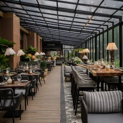 The dining space at FIORI, Lonavala, is designed to connect with its serene, verdant surroundings. A glass roof and walls create an al fresco feel, while the material palette of stone and wood-finished aluminum ensures durability and aligns with our sustainable practices.