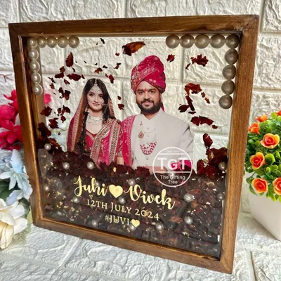 A stunning 10-inch square teakwood frame preserving a couple's varmala. The deep red flowers and pearls are set in clear resin, with a central photo and golden text for the names and date.