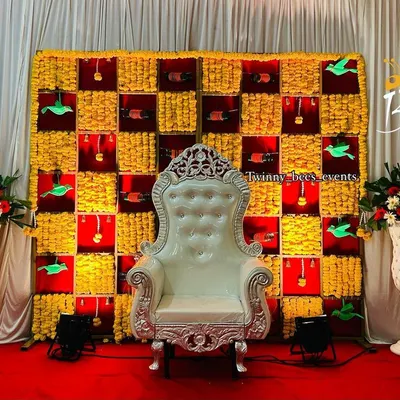 A vibrant and playful backdrop perfect for a baby shower or seemantham ceremony. The design features a checkerboard pattern of yellow marigolds and red fabric, with cute paper birds and bells hanging in the squares.