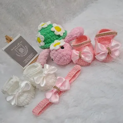 This baby gift hamper is filled with handmade treasures. It includes two pairs of booties with satin bows, one in white and one in pink, a matching pink headband, and a cute turtle toy with a daisy shell.