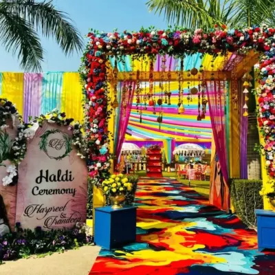 A complete Haldi ceremony entrance designed for maximum impact. The path is defined by a custom-painted floor, leading through a floral archway under a canopy of bright yellow, pink, and blue drapes.
