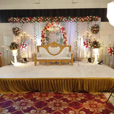 A classic and elegant stage setup for a wedding reception or engagement. It features a beautiful floral arch, a central chandelier, and warm candle-style lighting, all centered around a formal white and gold sofa for the couple.