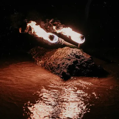 An atmospheric shot of two lit torches resting on a rock in the water at night, their flames reflected in the dark water. This sets the scene for a dramatic and fiery beach pre-wedding photoshoot.