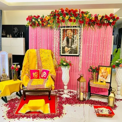 A full view of a Guruji satsang decoration I did in a client's living room. The setup includes the main seat, floral arrangements on stands, and a decorated portrait of Guruji.