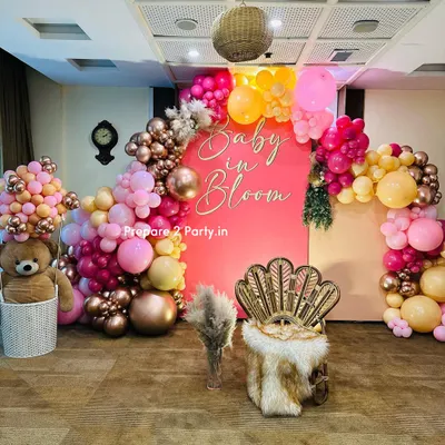 A vibrant and stylish "Baby in Bloom" themed baby shower. The setup features a bold pink backdrop, a stunning balloon arch in shades of pink, orange, and rose gold, and a chic peacock chair for the mother-to-be.