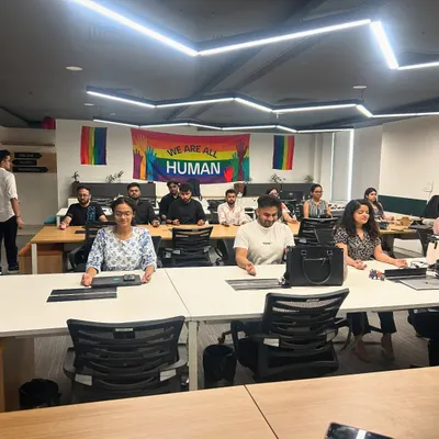 Leading a corporate yoga session at Zoo Media Network. The office is filled with a great vibe, with a "We Are All Human" pride flag in the background, reflecting a culture of inclusivity.