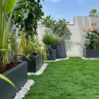 A closer look at a terrace makeover, where we've used a combination of rectangular planters and unique face-themed pots to add personality. The white pebbles create a clean border against the artificial grass, defining the green space.