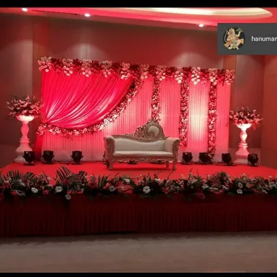 A classic and royal reception stage design. This setup features a beautiful contrast of red drapes and white flowers, with a regal sofa for the couple and elegant floral stands.